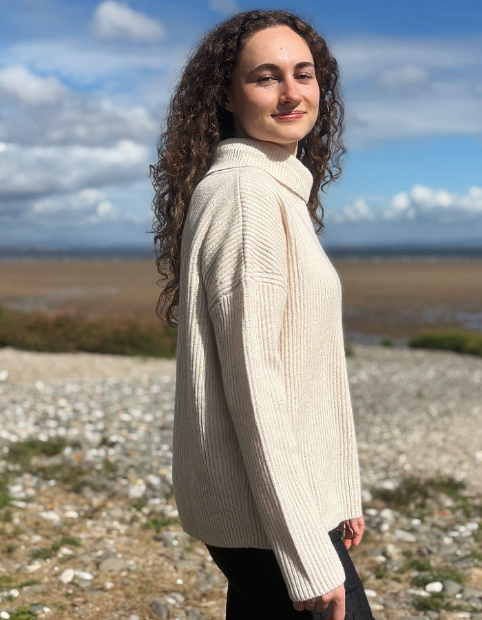 A woman with long curly brown hair stands on a rocky beach wearing a cream-colored ribbed turtleneck sweater and black pants, with a blue sky and sea in the background.