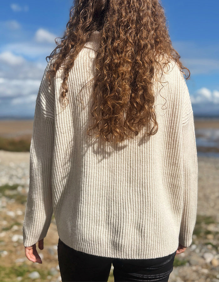 A person viewed from behind wearing a cream-colored ribbed knit sweater and black pants, standing outdoors on a rocky shoreline under a blue sky with clouds.