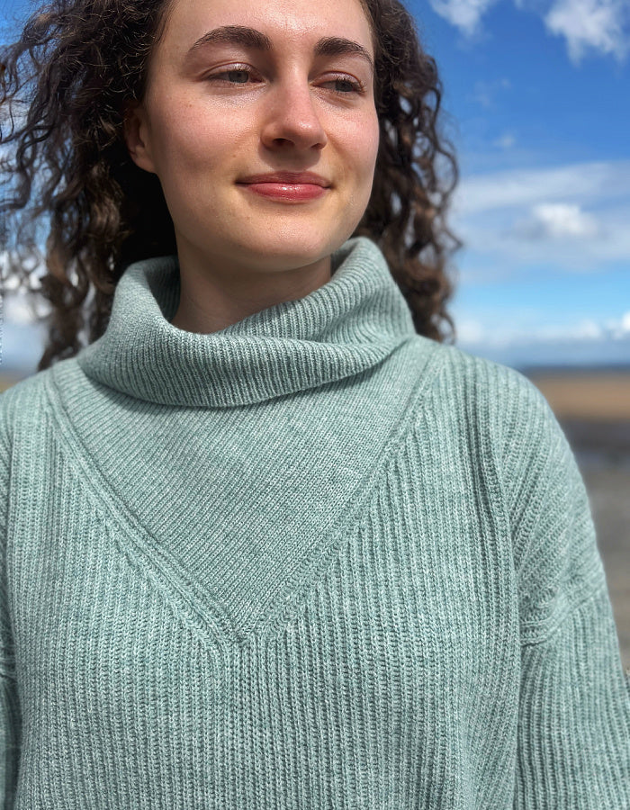 A close-up shot of a woman wearing a ribbed knit turtleneck sweater in a dusty sage green color, photographed outdoors against a blurred background of blue sky and water.