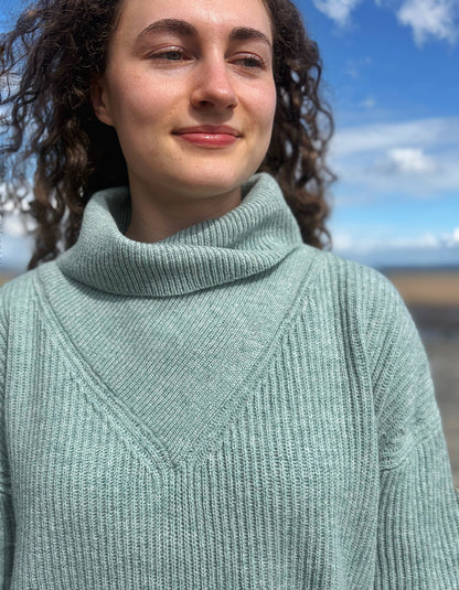 A close-up shot of a woman wearing a ribbed knit turtleneck sweater in a dusty sage green color, photographed outdoors against a blurred background of blue sky and water.