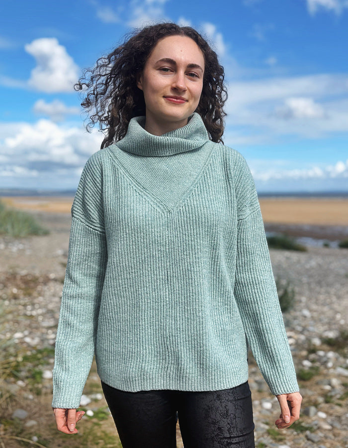 A woman with curly dark hair wearing a light sage green ribbed turtleneck sweater and black pants, standing outdoors on a rocky shoreline under a blue sky with clouds.