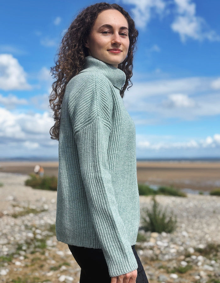 A woman with curly brown hair wearing a ribbed light sage green turtleneck sweater, standing outdoors on a rocky shoreline under a blue sky with clouds.