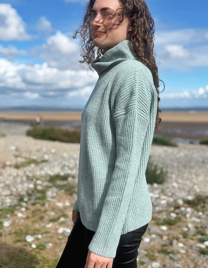 A woman with curly brown hair wearing a light sage green ribbed turtleneck sweater and black pants, standing outdoors on a rocky shoreline under a blue sky with clouds.