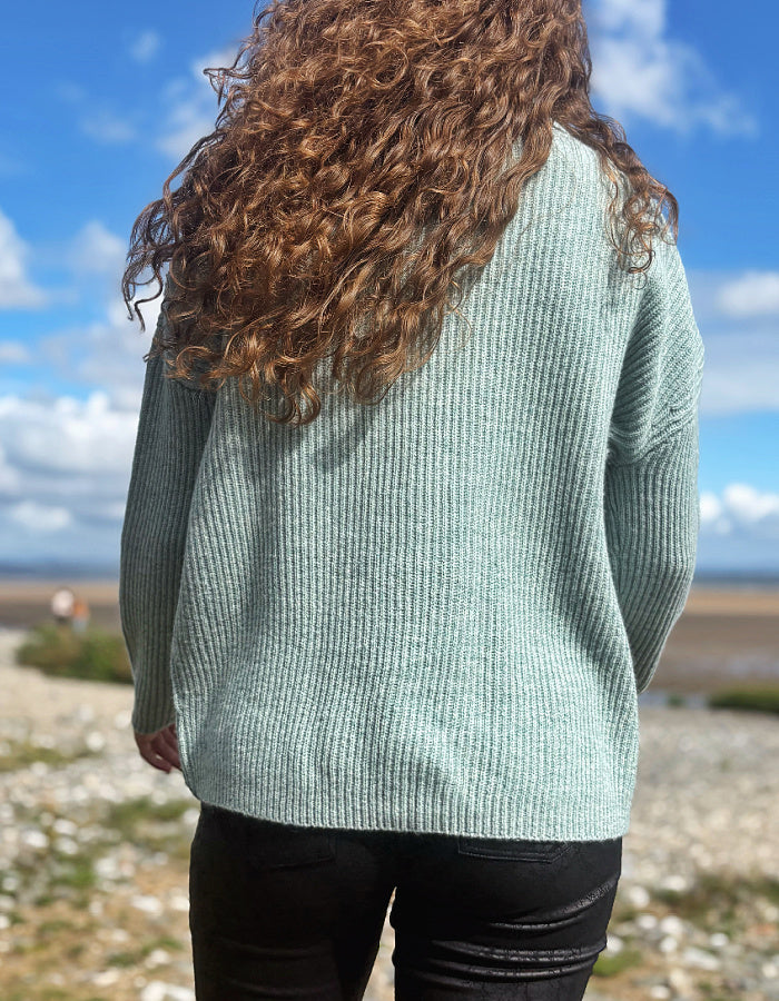 A person viewed from behind wearing a ribbed knit sweater in a pale sage green color, standing outdoors on a rocky beach under a blue sky with clouds.