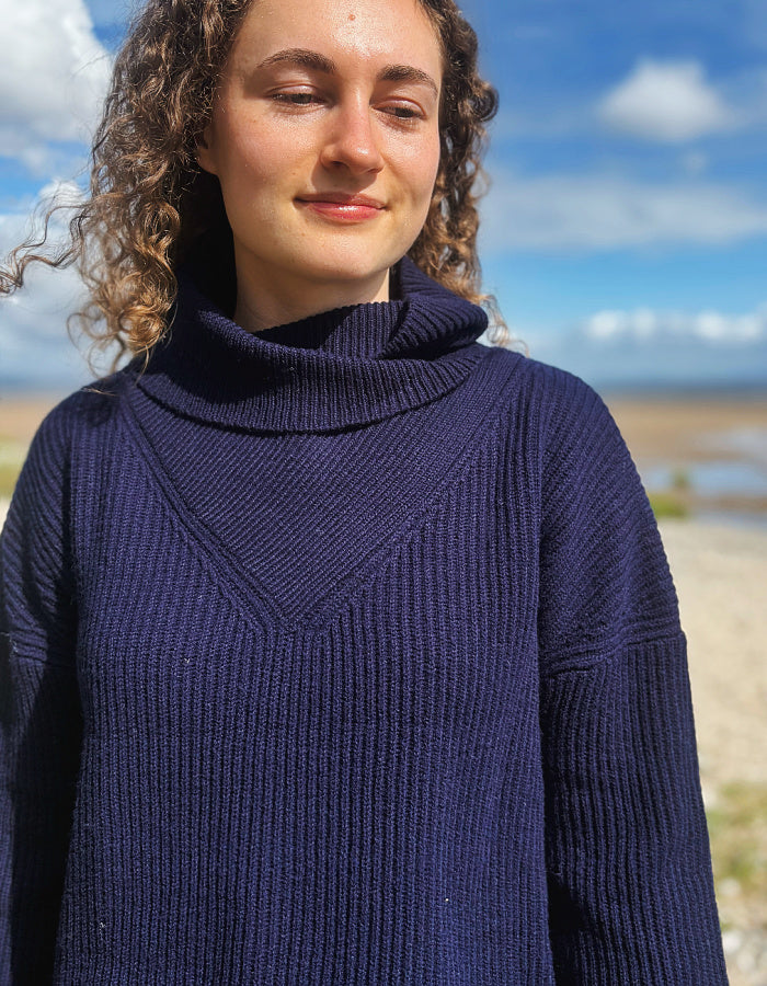 A woman with curly brown hair wearing a dark navy blue ribbed turtleneck sweater, photographed outdoors against a blurred background of blue sky and water.