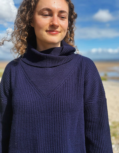 A woman with curly brown hair wearing a dark navy blue ribbed turtleneck sweater, photographed outdoors against a blurred background of blue sky and water.