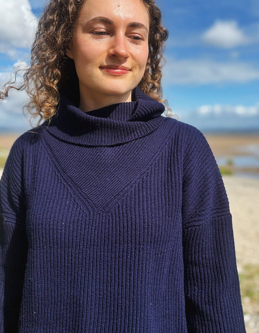 A woman with curly brown hair wearing a dark navy blue ribbed turtleneck sweater, photographed outdoors against a blurred background of blue sky and water.
