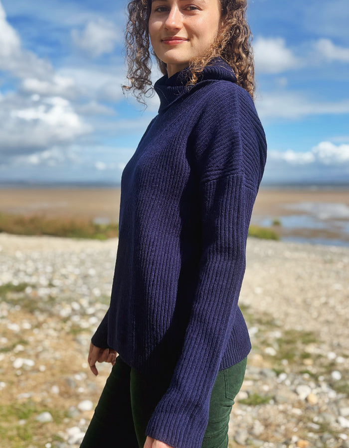 A woman wearing a dark navy ribbed turtleneck sweater and green pants, standing outdoors on a rocky beach with a cloudy blue sky in the background.
