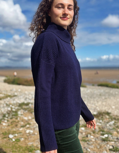 A woman with curly brown hair wearing a dark navy ribbed turtleneck sweater and green trousers, standing outdoors on a pebble beach with a blue sky and clouds in the background.