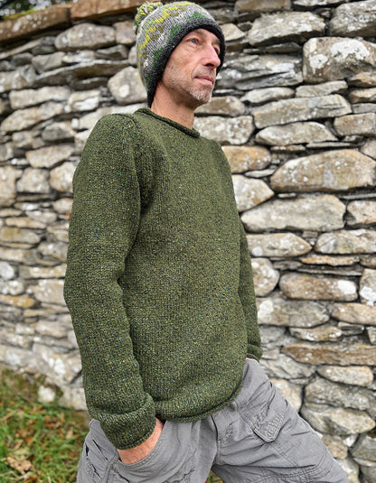 Man wearing a green knitted sweater and beanie standing against a stone wall.