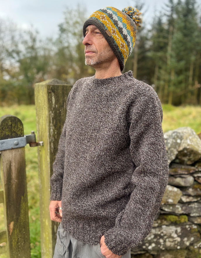 Man wearing a knitted sweater and hat standing by a wooden gate in a natural setting