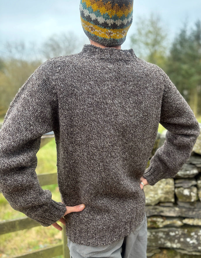 Person wearing a textured sweater and colorful knit hat outdoors with greenery and stone wall in the background.