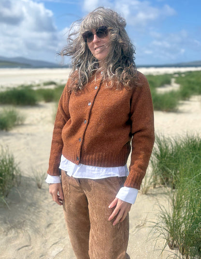 A woman standing outdoors on a sandy beach dune wearing a rust-orange knit cardigan over a white shirt and brown corduroy trousers, with grasses and blue sky in the background.