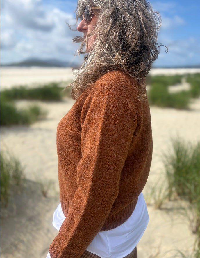 A woman standing outdoors on a sandy dune wearing a textured rust-orange knit sweater over a white top, with blurred beach grass and sky in the background.