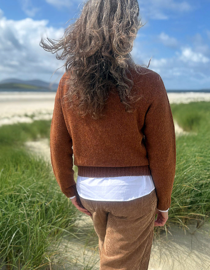 A person viewed from behind wearing a rust-brown knit sweater, a white shirt, and brown corduroy trousers, standing on a sandy path in a grassy coastal area under a blue sky.