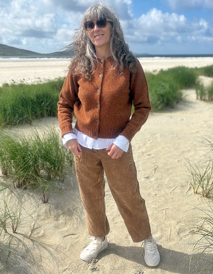 A woman stands on a sandy beach dune wearing a brown textured button-up cardigan over a white shirt, brown corduroy trousers, and white sneakers, with a beach and blue sky in the background.