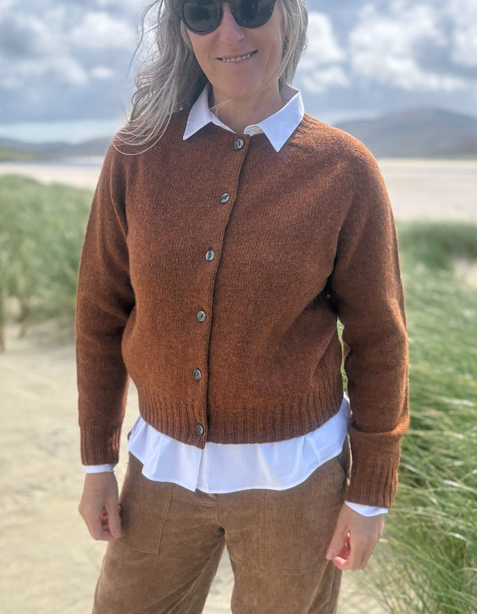 A woman wearing a brown knit button-up cardigan over a white collared shirt, standing outdoors on a sandy path with grass and a cloudy sky in the background.