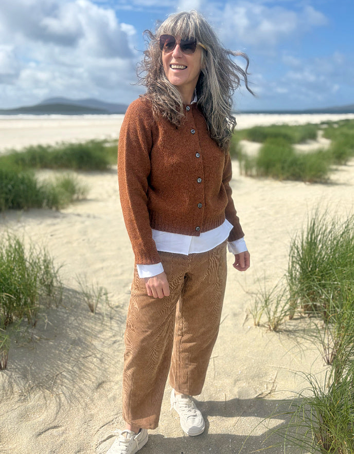 A woman standing on a sandy beach wearing a brown textured cardigan over a white shirt, brown corduroy pants, and white sneakers, with dunes, grass, and a cloudy blue sky in the background.