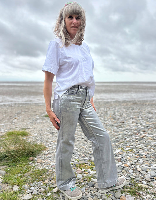 Person wearing a white t-shirt and light silver wide leg jeans standing on a pebbly beach with a cloudy sky.