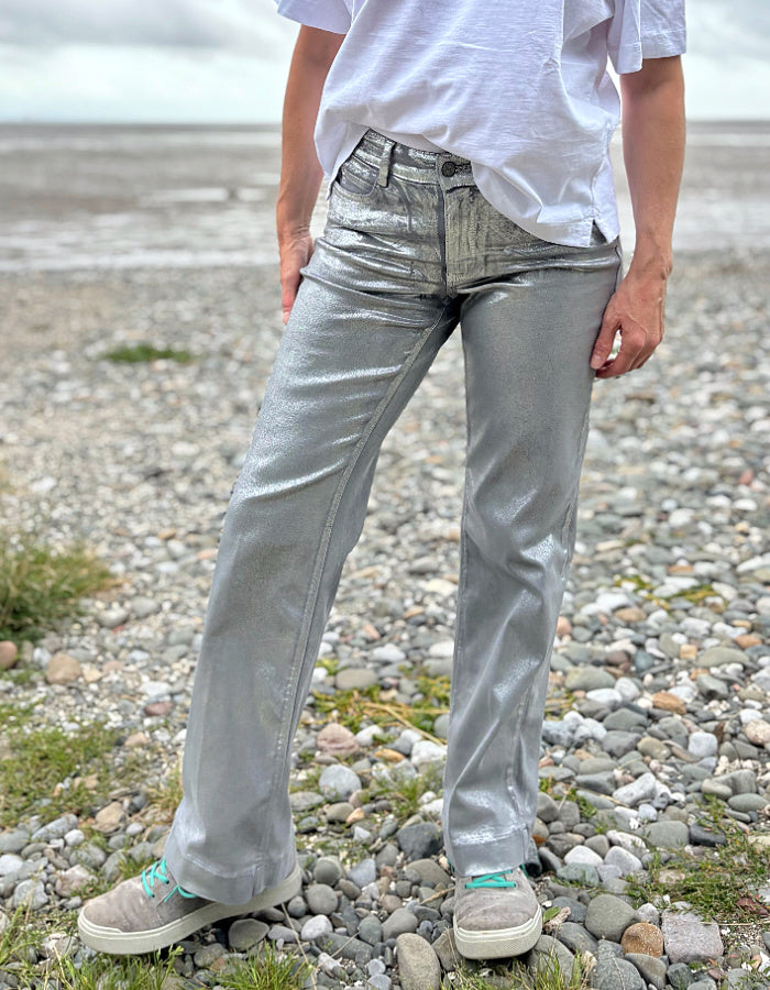 Person wearing a white t-shirt and light silver wide leg jeans standing on a pebbly beach with a cloudy sky.