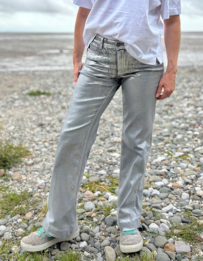 Person wearing a white t-shirt and light silver wide leg jeans standing on a pebbly beach with a cloudy sky.