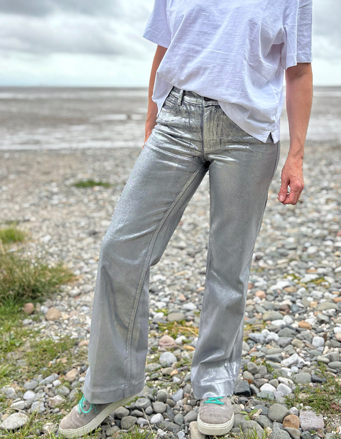 Person wearing a white t-shirt and light silver wide leg jeans standing on a pebbly beach with a cloudy sky.