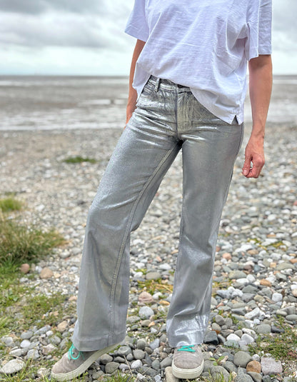 Person wearing a white t-shirt and light silver wide leg jeans standing on a pebbly beach with a cloudy sky.