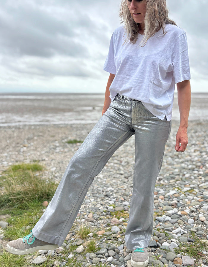 Person wearing a white t-shirt and light silver wide leg jeans standing on a pebbly beach with a cloudy sky.