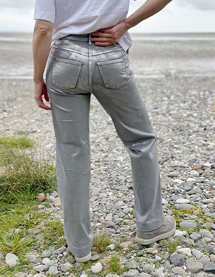 Person wearing a white t-shirt and light silver wide leg jeans standing on a pebbly beach with a cloudy sky.