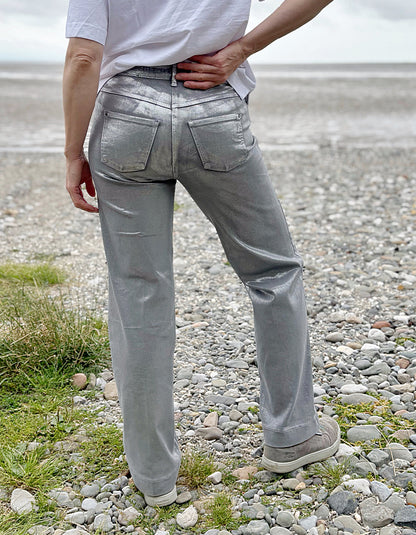 Person wearing a white t-shirt and light silver wide leg jeans standing on a pebbly beach with a cloudy sky.