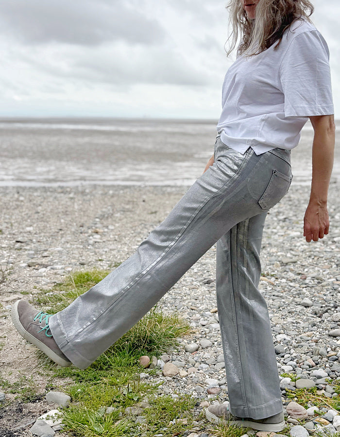 Person wearing a white t-shirt and light silver wide leg jeans standing on a pebbly beach with a cloudy sky.