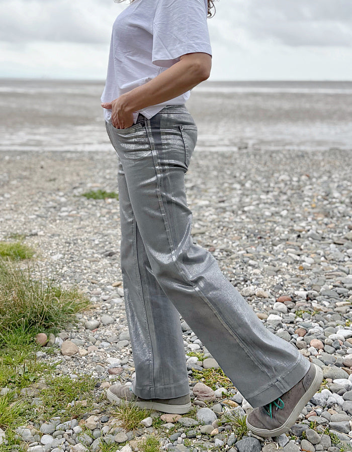 Person wearing a white t-shirt and light silver wide leg jeans standing on a pebbly beach with a cloudy sky.