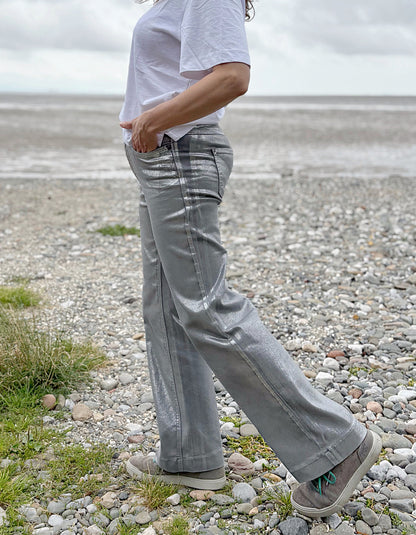 Person wearing a white t-shirt and light silver wide leg jeans standing on a pebbly beach with a cloudy sky.