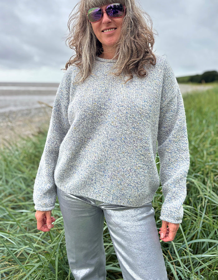 Person wearing a gray sweater and sunglasses standing in a grassy field with a beach in the background.