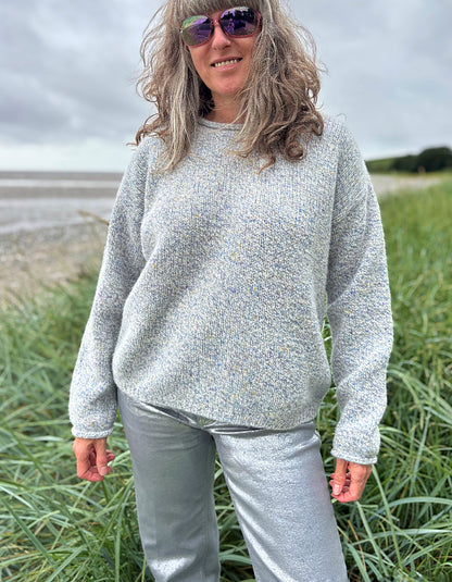 Person wearing a gray sweater and sunglasses standing in a grassy field with a beach in the background.