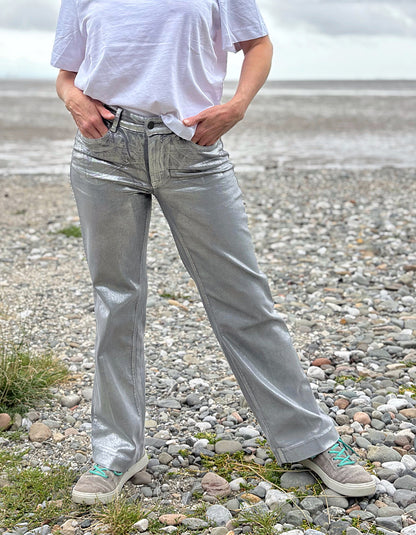 Person wearing a white t-shirt and light silver wide leg jeans standing on a pebbly beach with a cloudy sky.