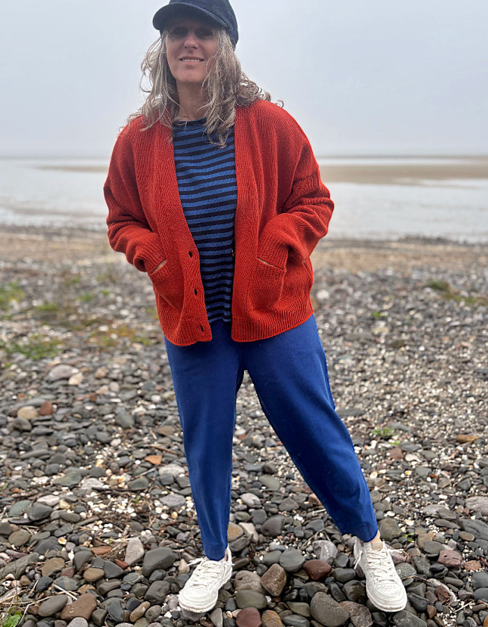 A woman stands on a pebble beach wearing a rust-red cardigan over a navy and blue striped shirt, blue pants, white sneakers, and a black cap.