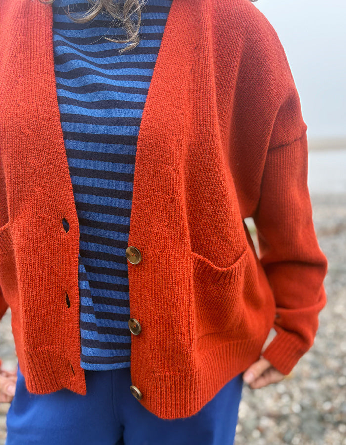A person wearing a rust-orange knit cardigan over a blue and navy striped top, paired with blue pants, photographed outdoors on a pebble beach.