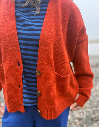 A person wearing a rust-orange knit cardigan over a blue and navy striped top, paired with blue pants, photographed outdoors on a pebble beach.