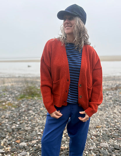 A woman standing on a pebble beach wearing a rust-orange knit cardigan over a blue striped shirt, blue pants, and a dark navy cap.