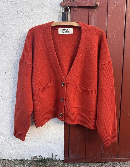 A rust-orange knit cardigan sweater hanging on a wooden hanger against a split white stucco wall and dark red wooden door. The cardigan features a V-neck, three front buttons, dropped shoulders, and two front patch pockets.