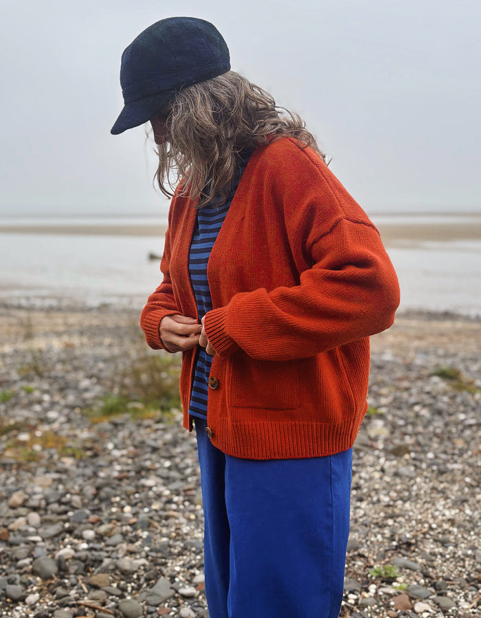 A person standing on a pebble beach wearing a rust-orange knit cardigan over a blue striped top and blue pants, with a dark navy cap in hand. The background shows a muted, overcast shoreline.