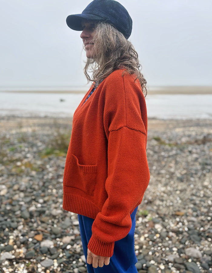 A woman standing on a pebble beach wearing a bright rust-orange knit sweater, blue pants, and a dark navy cap, shown in profile.