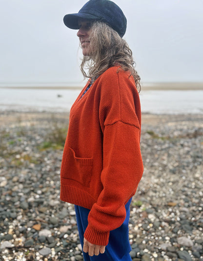 A woman standing on a pebble beach wearing a bright rust-orange knit sweater, blue pants, and a dark navy cap, shown in profile.