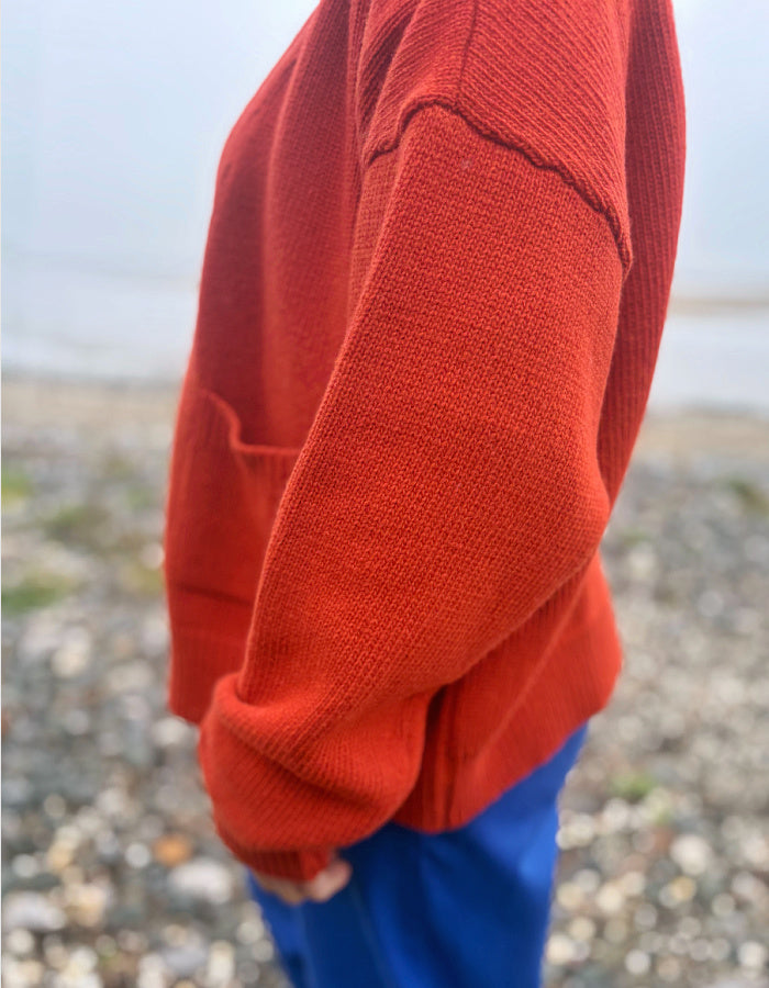 A close-up side view of a person wearing a textured rust-orange knit sweater and blue pants, standing on a pebble beach with a blurred background.