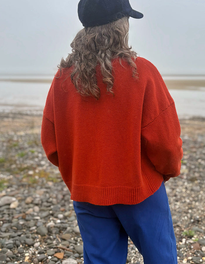 A person standing on a pebble beach, viewed from behind, wearing a bright rust-orange knit sweater, blue pants, and a black cap.