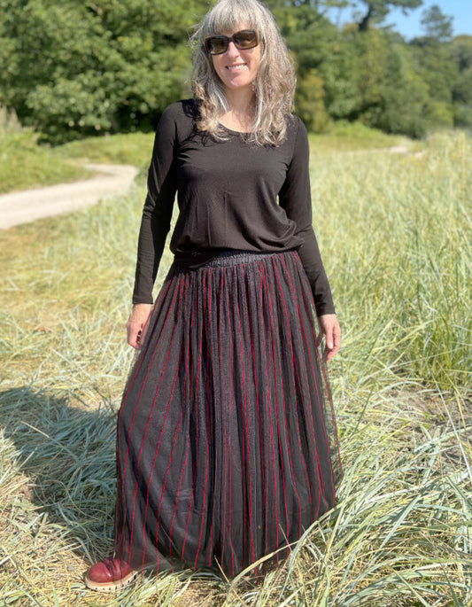 Woman standing in a field wearing a black long-sleeve top and a long, maxi length dark skirt with red stripes.