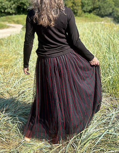 Woman standing in a field wearing a black long-sleeve top and a long, maxi length dark skirt with red stripes.