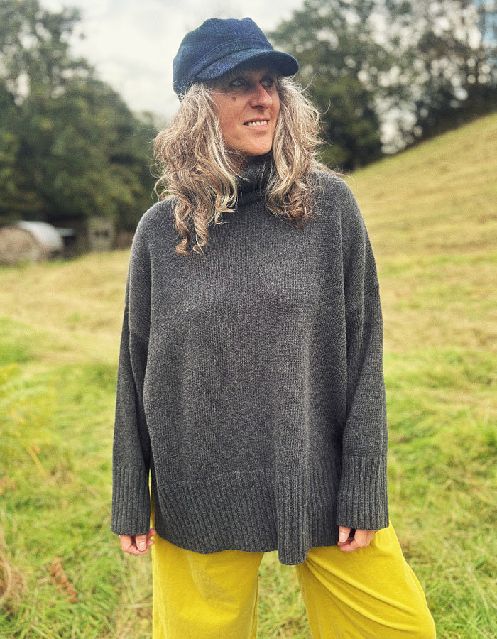A woman wearing an oversized dark gray turtleneck sweater, a navy blue cap, and mustard yellow pants, standing in a grassy outdoor field.