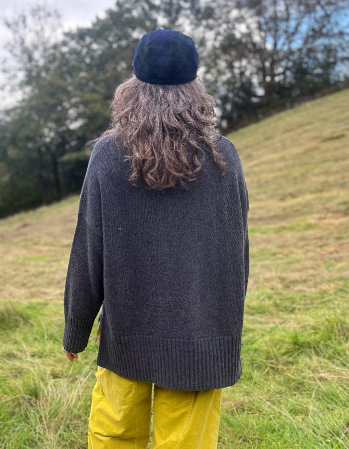 A person standing outdoors on a grassy hill, viewed from behind, wearing a dark charcoal gray knit sweater, a dark navy beret, and bright yellow pants.